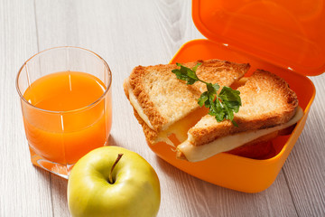 Yellow lunch box with toasted slices of bread, cheese and green parsley, green apple and glass of orange juice on wooden desk.