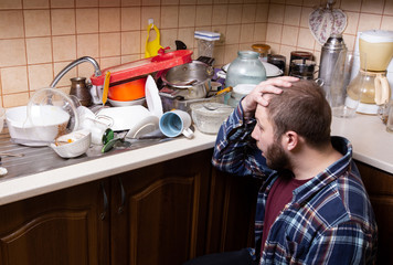 A young bearded guy sits on the floor and is shocked by the amount of dirty dishes lying in the kitchen sink to be washed.