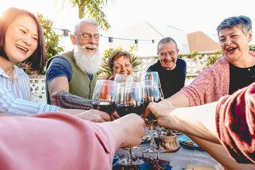 Happy senior friends toasting with red wine glasses at dinner on patio - Mature people having fun dining together outside - Elderly lifestyle, food and drink, retired and pensioners concept