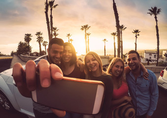 Happy friends standing in front of convertible car and taking selfie with mobile phone camera -...