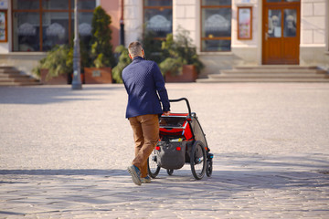 A young man with a child in a modern tourist stroller walking through the city center. The responsibility of the father for the upbringing of the younger generation. Family vacation. Care of offspring