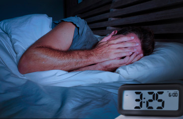  sad man awake lying sleepless on bed covering his eyes crying suffering insomnia sleeping disorder with alarm clock - obrazy, fototapety, plakaty