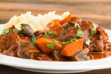 Coq au Vin, traditional French recipe of chicken braised in red wine with bacon, carrot and mushrooms. Served with mashed potatoes on white plate. Macro