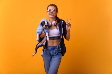 Fototapeta na wymiar High fashion look.glamor stylish beautiful young woman model.Attractive, charming, pretty girl in stylish outlook, holding french bulldog on arms, looking at camera, standing over yellow background 