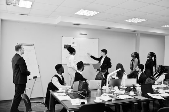 Mixed race business coach presenting report standing near whiteboard pointing on sales statistic shown on diagram and chart teach diverse company members gathered together in conference room.