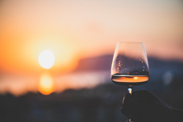Man's hand holding glass of rose wine and with sea and beautiful sunset at background, close-up,...