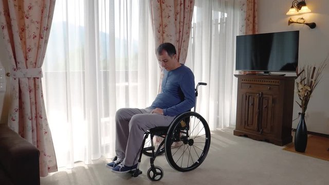 Sad young man in wheelchair looking through the window at home or in nursing home. Slow motion 4k hand held movement