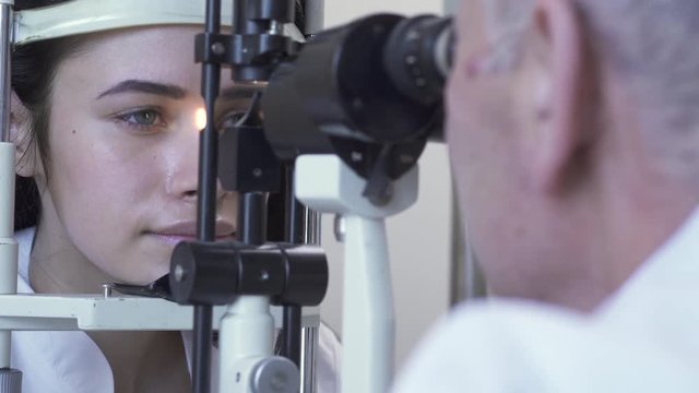 Pretty caucasian woman do eye test at clinic sitting in front of doctor in white medical coat looking in eye test machine. Mature optometrist examining patient eye using light. Health care