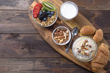 full Breakfast of oatmeal with nuts, muesli with berries and fruits, croissants and glass of milk on wooden background