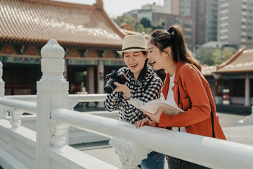 happy young best friends checks photos on camera standing by white marble stone railing in Chinese...