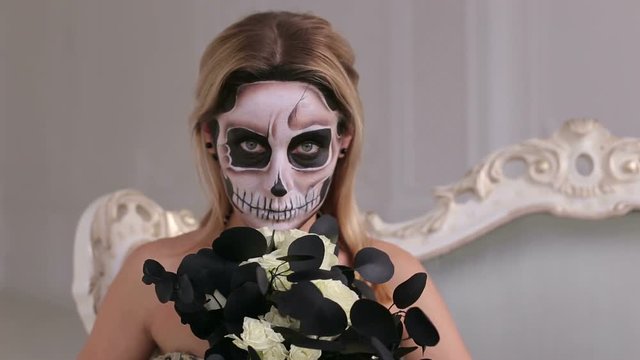 A young blonde in a black dress with a bouquet of black flowers and makeup in the form of a skeleton, in the image of a witch Halloween posing in a bright room with a mirror wall.