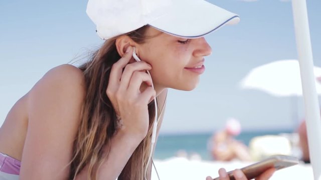 beautiful tanned happy laughing woman is lying on the beach on a white sand, relaxing and listen music from the phone with small headphones. slim sexy woman in bikini and white cap enjoing and