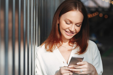 Plakat Pleased woman focused into screen of cell phone, checks email box, dressed in white clothes, sends feedback, connected to wireless internet, has brown hair, charming smile, sends text message