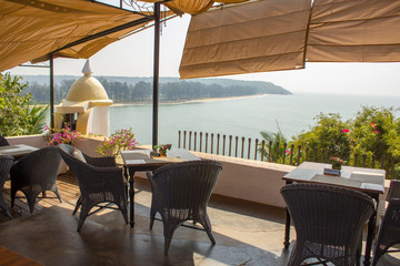 empty tables and chairs in a restaurant against the backdrop of the sea and the coast with green trees