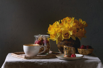Layered dessert in jar. Biscuit, coffee cream, nuts, raspberries, fresh mint. Served with cup of coffee espresso, berries, daffodils flowers and jug of milk over grey linen table cloth.