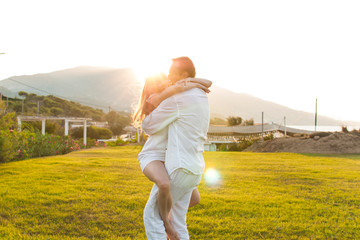 Romantic view of happy couple in white clothes, when they cuddle on green grass. Beautiful landscape of sun above mountain during sunset. Concept of honey moon love.