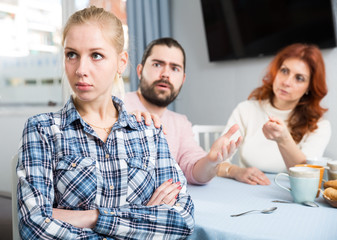 Upset girl sitting  at table after quarrel and man with mother on background