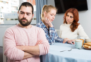 Upset young man sitting  at table after quarrel and girl with mother on background