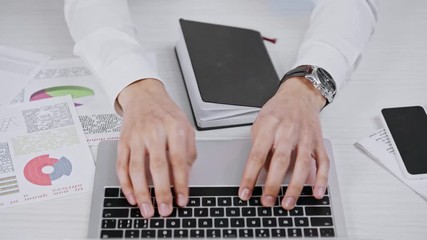 cropped view of businessman using laptop and writing in notebook with pen in office