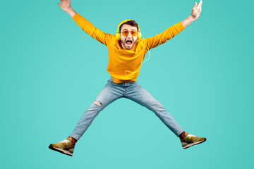 Fototapeta na wymiar Crazy funny bearded young man wearing yellow sunglasses listening to music with yellow headphones jump over cyan background.