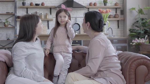 Grandmother, mother and little daughter spend time together sitting on the sofa in studio apartment. The girl standing on the cout, mom and granny looking at her. Child is smiling.