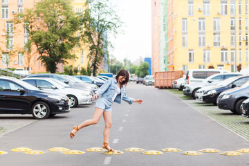 funny happy young woman in blue denim casual style jumping and walking on yellow road city bump on summertime day. city background on summer day.