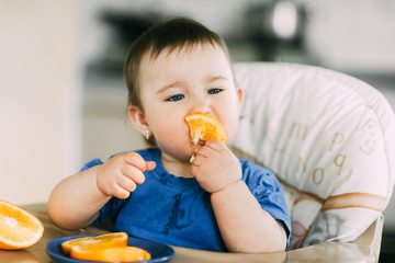 little girl sitting in baby chair, eating an orange