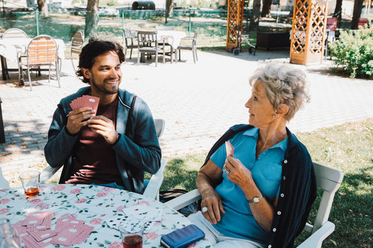 Retired senior woman playing cards with smiling male caretaker sitting at table in back yard