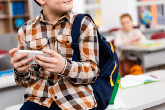 Partial view of pupil in checkered shirt with backpack using smartphone in classroom