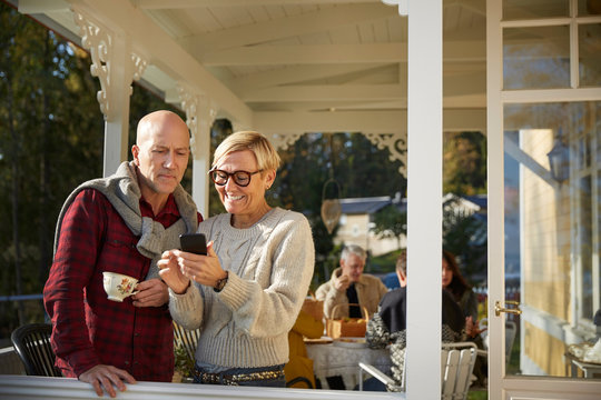 Cheerful mature woman showing mobile phone to bald man with friends in background on porch
