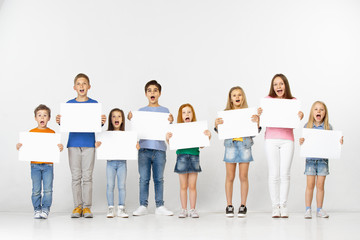 Fototapeta premium Group of happy surprised screaming children with a white empty banners isolated in light studio background. Education and advertising concept