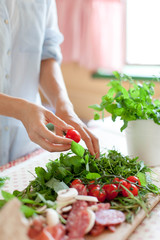 Woman is cooking in home kitchen. Female hands choose cherry tomatoes and vegetables. Ingredients for preparing italian or french food are on table on wooden boards. Lifestyle moment. Close up.