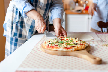 Obraz na płótnie Canvas Woman cuts hot pizza on wooden board in cozy home kitchen. Cooking process of italian family dinner at home. Lifestyle moment. Close up of female hands with knife.