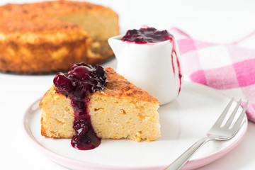 One slice of millet cake with berry sacue on white plate. Whole cake and table cloth in the background.