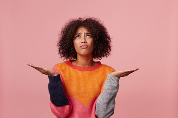 Brunette girl with afro hairstyle offended sad upset, in bad mood, stand with palms raised up holding a copy space, feels injustice hopelessness wearing colorful sweater, isolated on pink