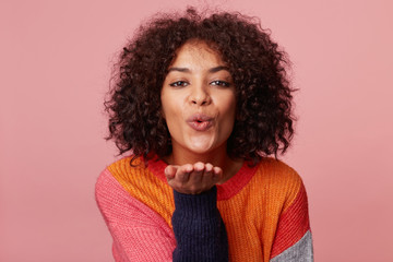 Close up portrait of charming african american girl with afro hairstyle blowing air kiss from open palm,expresses her devotion and truthful love to boyfriend, isolated on pink background