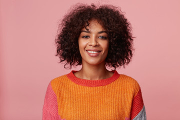Close up portrait of positive optimistic charming african american girl with afro hairstyle looks with pleasure, with friendly smile, wearing colorful longsleeve, isolated on pink background