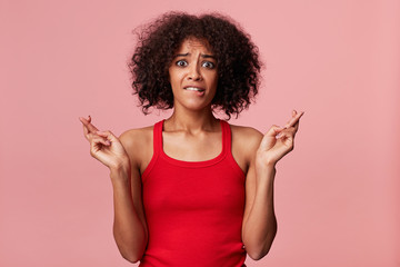 Attractive young african american woman nervous, feels panic, crossed her fingers and bites her lip while worries about something, waiting for a call, positive news, results,isolated on pink backgroud