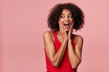 Young attractive African American with an afro hairstyle with delight looks left to the empty space, her hands touch her face, feels pleased, looks surprised, isolated on a pink background