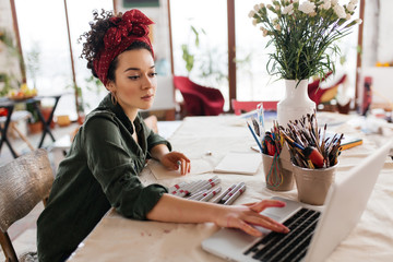 Young beautiful woman with dark curly hair sitting at the table dreamily using laptop and drawing...