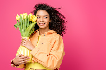 attractive smiling african american girl holding vase with yellow tulips while standing isolated on crimson