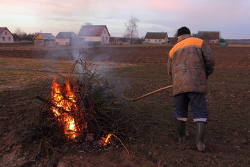 Spring cleaning in the backyard - a male farmer in overalls burns dry branches at the edge of the...