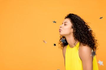 happy curly african american girl in yellow dress blowing near shiny confetti stars isolated on...