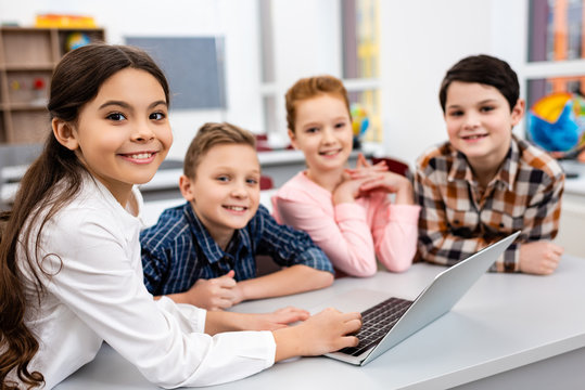 Happy preteen pupils using laptop in classroom with smile