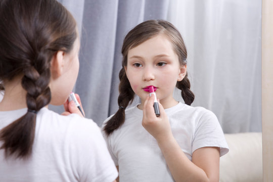 A little girl looks in the mirror and paints her lips with bright lipstick.