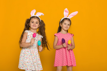 two little girls with Easter bunny ears holding colorful eggs on yellow background. Happy easter. cute children