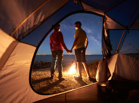 View from inside tourist tent. Young romantic couple, man and woman standing at campfire holding hands on lake shore under blue starry evening sky on lake water background. Tourism and love concept.