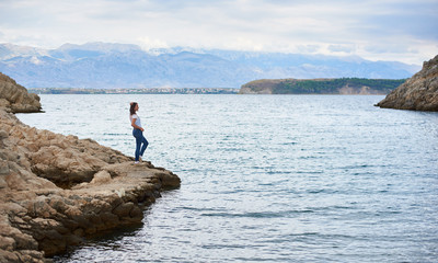 Fototapeta na wymiar Young attractive slim woman in jeans and T-shirt standing alone on stony shore of sea, enjoying beautiful view. Clear shiny water and mountains on background. Tourism and vacations concept.