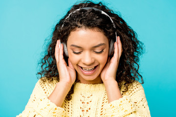 attractive curly african american girl listening music in headphones with closed eyes isolated on blue