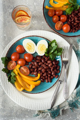 Healthy food, red beans, tomatoes, boiled egg, pepper with parsley in a plate with devices and a glass of fruit water on a gray background top view.